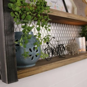May include: Two-tiered metal and wood shelf with a geometric pattern on the front. The shelf is decorated with a blue ceramic planter with a plant, two small metal bicycles, and a silver metal sheep. The shelf is mounted on a white wall.