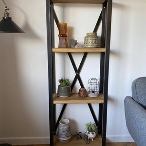 May include: A black metal shelf unit with four wooden shelves. The shelves are decorated with various items, including vases, plants, and decorative objects. The shelf unit is standing on a wooden floor.