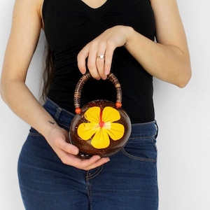 May include: A round, dark brown coconut shell handbag featuring a bright yellow five-petal flower design on the front. The bag has a curved, textured handle made of dark brown material with small orange beads at the base of the handle attachment.