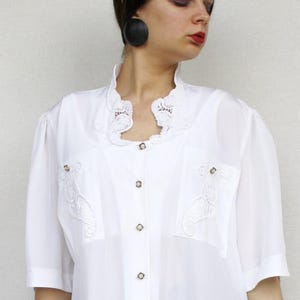 Può includere: Camicetta bianca con maniche corte, colletto alto con finiture in pizzo e ricami decorativi sulle tasche. La camicetta ha bottoni color oro sul davanti. La modella indossa jeans blu.