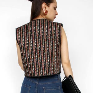 Puede incluir: Un chaleco sin mangas con base negra y rayas verticales de patrones florales en rojo, blanco y dorado, combinado con vaqueros azules, botas negras hasta la rodilla y un bolso de mano negro. El chaleco tiene un borde negro en los acabados.