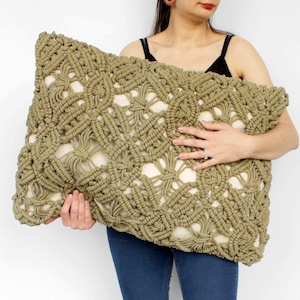 May include: A large, rectangular macrame pillow in shades of olive green and beige. The pillow features a detailed, woven pattern. The pillow is held in front of a white background.