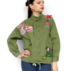 Könnte beinhalten: Olivgrüne Jacke mit Blumen- und Patch-Verzierungen. Die Jacke hat einen Stehkragen, Vordertaschen und eine Kordelzug-Taille. Das Model trägt blaue Jeans und schwarze Stiefel.