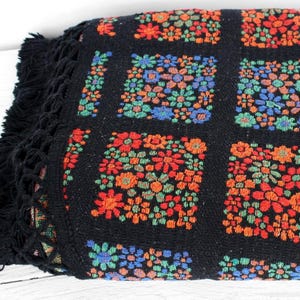 May include: A folded black blanket with a floral pattern in red, orange, blue, and green. The blanket has a grid design with each square filled with flowers. The edge has a black fringe.