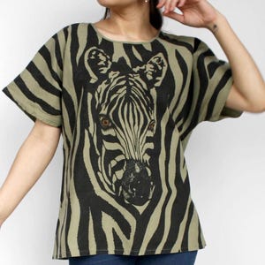 May include: Olive green and black zebra print top. The short-sleeved shirt features a large zebra face graphic on the front. The model is wearing blue jeans. The top has a relaxed fit.