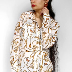 May include: White button-down shirt with a gold chain and butterfly pattern. The long-sleeved shirt has a collared neckline and two chest pockets. The shirt is paired with dark blue jeans.