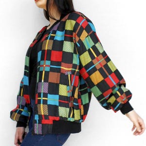 Peut inclure: Veste de style bomber colorée avec une base noire et un motif à carreaux vibrant en rouge, jaune, bleu et vert. La veste a un col et des poignets noirs, et est associée à un jean bleu.