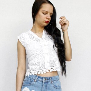 Puede incluir: Un top sin mangas de ganchillo blanco con cuello en V, cierre frontal de botones pequeños blancos y dobladillo festoneado, combinado con pantalones cortos de mezclilla azul claro con roturas y dobladillo deshilachado.
