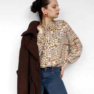 May include: A long-sleeved button-down shirt with a cream base and a geometric pattern in red, green, and gold, paired with dark blue denim jeans, a dark brown wool coat, and brown leather lace-up shoes with square-shaped red earrings.