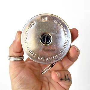 May include: A silver-colored tape measure with black text and a black center button. The tape measure has a metal clip and is held in a hand. The text includes "ГОСТ 7502-80" and "3ИПК5-10ДAYT/1".