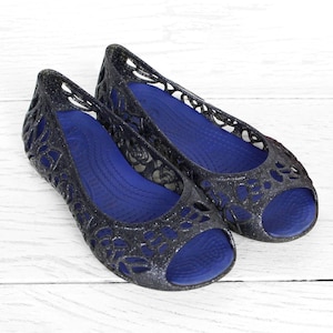May include: A pair of black, open-toe ballet flats with a glittery finish. The shoes have a floral cutout design on the upper and a bright blue insole. The flats are displayed on a white wooden surface.