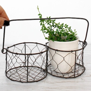 Könnte beinhalten: Ein rustikaler Drahtkorb mit zwei Fächern und einem Griff, der einen kleinen weißen Keramiktopf mit einer grünen Pflanze enthält. Der Korb hat eine dunkelbraune Oberfläche und steht auf einer hellen Holzoberfläche.