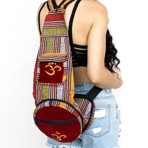 Op de afbeelding: Een kleurrijke rugzak met een rood, geel en blauw patroon. De rugzak heeft een rode zak met een gouden Om-symbool. De tas heeft een ritssluiting en verstelbare bandjes.