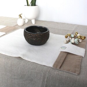 May include: Two rectangular placemats featuring a white center panel and a tan border on one side, secured with a single button. A dark brown textured bowl sits on one mat, while a small decorative branch with light-colored beads rests on the other.