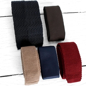 May include: Six knitted ties in a range of colours, such as black, brown, beige, navy, and burgundy, are arranged on a white wooden surface. The ties have a textured finish and are neatly displayed, highlighting their different shades and patterns.