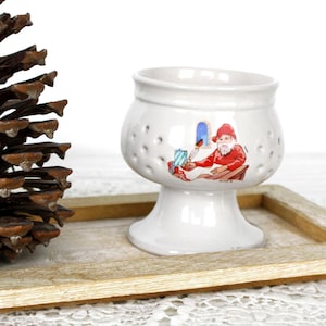 May include: A white ceramic egg cup featuring a small illustration of a bearded figure in a red hat writing at a desk. The cup has a flared base and a series of small circular perforations around the upper bowl section, providing a decorative detail.