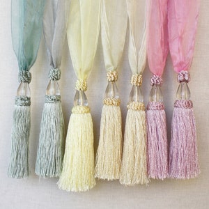 May include: A selection of decorative tassels in a row, each with a different pastel hue. The tassels are attached to clear, teardrop-shaped ornaments. The colours include shades of blue, green, yellow, and pink. The background is a neutral, textured fabric.