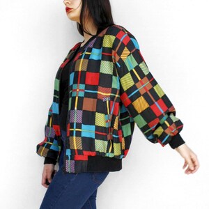 May include: A colorful, vintage bomber jacket with a black base and a vibrant plaid pattern in red, yellow, blue, and green. The jacket has a black collar and cuffs, and a zipper closure. The sleeves are long and slightly puffed.
