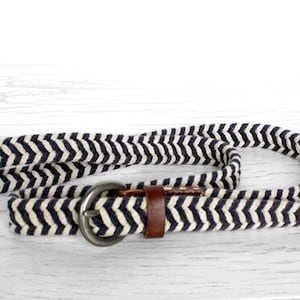 Op de afbeelding: Een marineblauwe en witte chevron patroon riem met een zilveren gesp en bruine lederen accenten. De riem is gemaakt van een geweven materiaal en ligt op een wit houten oppervlak.