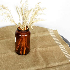 May include: A brown glass jar filled with dried wheat stalks sits on a tan linen tablecloth with white stripes. The jar is cylindrical with a wide mouth. The tablecloth is draped over a dark, round surface.