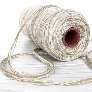 Puede incluir: Una bobina de hilo de color natural con un núcleo marrón. El hilo está enrollado firmemente alrededor del núcleo, con una longitud del hilo desenrollada y en bucle sobre una superficie blanca. El hilo tiene un aspecto texturizado.