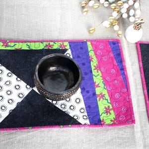 May include: A vibrant quilted placemat with a geometric design. The placemat has a black and white triangle pattern, a pink border, and vertical stripes in purple, green, and pink. A dark bowl rests on the placemat.