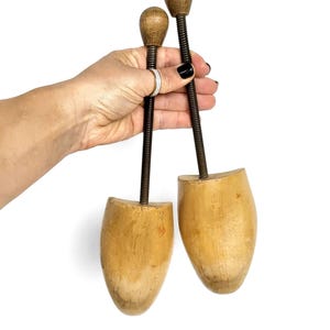 May include: A pair of vintage wooden shoe stretchers, featuring metal rods and wooden knobs. The shoe forms are light brown, with a classic design. The metal rods are dark brown. The shoe stretchers are held against a white background.