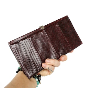 Op de afbeelding: Een rechthoekige, donkerbordeauxkleurige portemonnee met een gestructureerd oppervlak dat doet denken aan slangenleer. De portemonnee heeft een slanke, langwerpige vorm en is voorzien van een zilverkleurige metalen sluiting aan de bovenrand.