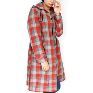 May include: A knee-length coat featuring a red and gray plaid pattern, a hood, long sleeves with buttoned cuffs, and a front button closure. The coat is worn over blue jeans and black platform boots, with a large black circular earring visible.