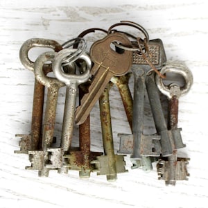 Op de afbeelding: Een verzameling antieke sleutels van verschillende maten en vormen, samengebonden met metalen ringen en kettingen. De sleutels vertonen tekenen van ouderdom met roest en slijtage, wat suggereert dat het vintage of hergebruikte items zijn. De achtergrond is een wit, getextureerd oppervlak.