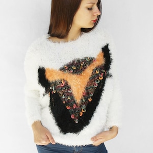 May include: A white, fluffy sweater with a black and orange geometric design on the front. The design is embellished with colorful sequins. The sweater is paired with blue jeans.