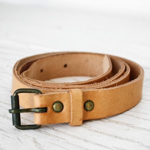 Puede incluir: Un cinturón de cuero marrón claro con una hebilla de latón. El cinturón está enrollado y la hebilla es visible.