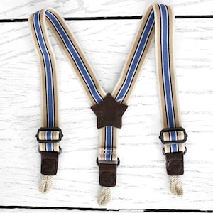 Peut inclure: Bretelles rayées avec des rayures bleues, beiges et noires. Les bretelles ont des détails en cuir marron et une pièce centrale en forme d'étoile marron. Elles sont conçues pour se fixer aux pantalons avec des clips et des boucles.