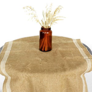 May include: A brown glass jar filled with dried wheat stalks sits on a tan table runner with three white stripes on each side. The table runner is draped over a dark gray table.