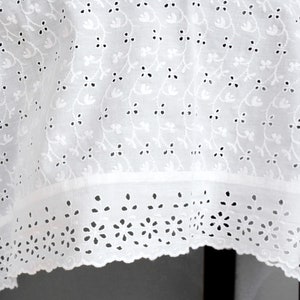 May include: A white fabric textile featuring an intricate eyelet embroidery pattern with small floral motifs and vines. The bottom edge is finished with a wide, scalloped border containing larger flower cutouts and decorative eyelet detailing.