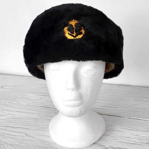 May include: Black faux fur hat with a gold embroidered emblem. The hat is displayed on a white mannequin head. The hat has a rounded shape and a soft texture. The background is light wood.