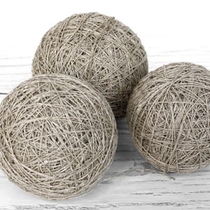 Può includere: Tre sfere di spago color naturale sono disposte su una superficie bianca e strutturata. Lo spago è avvolto strettamente, creando una superficie strutturata. Le sfere sono di dimensioni variabili, suggerendo un tema artigianale o decorativo.