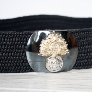 Puede incluir: Cinturón negro tejido con hebilla plateada y emblema dorado y plateado. El cinturón ancho tiene un aspecto texturizado. La hebilla es el elemento principal, añadiendo un toque decorativo al accesorio.