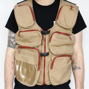 May include: Tan utility vest with multiple pockets and red zipper accents. The vest has a clear pocket and black buckles. A practical garment for outdoor activities or work.