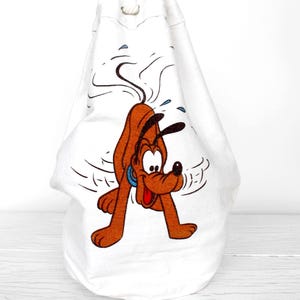 May include: A white drawstring bag featuring a graphic print of a brown dog with black ears, a blue collar, and a red tongue. The dog is depicted in a shaking motion with small blue water droplets and black motion lines surrounding the figure.