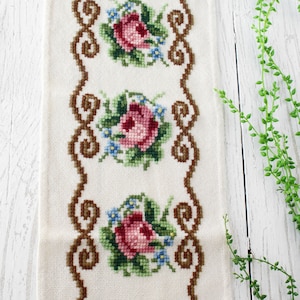 Può includere: Un runner da tavola rettangolare in tessuto color avorio con un motivo a punto croce di quattro composizioni floreali con rose rosa e piccoli fiori blu, racchiuso da un bordo a volute marrone e rifinito con frange sul lato superiore.