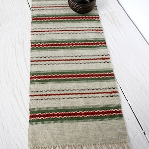 May include: A rectangular woven table runner featuring a light beige base with horizontal stripes in forest green and deep red, accented by small black geometric patterns. The runner has fringed ends and a small, dark brown textured bowl on top.