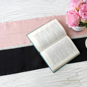 May include: A table runner with a pink and black color block design, accented with white lace trim. An open book, a bouquet of pink flowers, and a white candle holder are arranged on the runner, creating a decorative display.