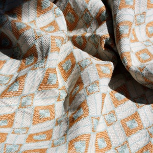 May include: A woven textile fabric featuring a repeating geometric pattern of diamonds and squares in shades of burnt orange, light blue, and off-white. The material is draped in soft folds, highlighting the textured weave and the intricate grid design.