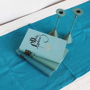 Peut inclure: Trois livres bleu clair avec des lettres dorées sont empilés sur un chemin de table turquoise. Deux bougeoirs turquoise avec des accents dorés sont également présents, créant une esthétique vintage.