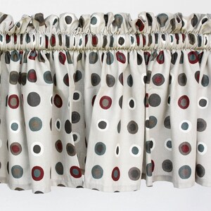 Può includere: Una mantovana per finestra in tessuto beige con un motivo ripetuto di punti circolari nelle tonalità grigio scuro, rosso intenso e bianco. La mantovana presenta un'intestazione arricciata con tasca per bastone, creando pieghe verticali.