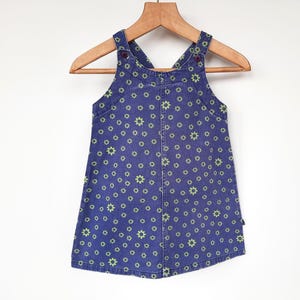 Op de afbeelding: Een blauwe spijkerbroekjurk met een groen bloemenpatroon. De mouwloze jurk heeft verstelbare bandjes en hangt aan een houten hanger. De jurk is ontworpen voor kinderen.