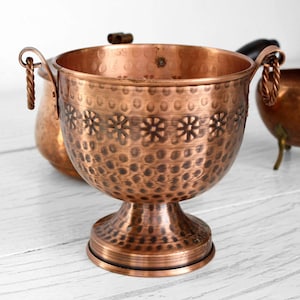 May include: A copper-colored, footed bowl with a textured surface and floral design. The bowl has two handles with a twisted rope design. Other copper-colored vessels are in the background.