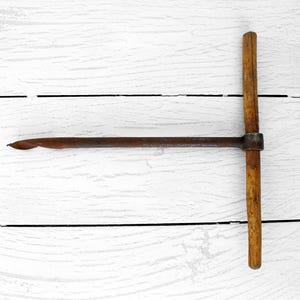 Puede incluir: Una herramienta antigua de metal oxidado y madera, con un mango en forma de T. La herramienta tiene un eje largo y delgado de metal oxidado con un extremo puntiagudo. El mango es de madera y está unido al eje con un accesorio de metal. El número "#1" está en la esquina inferior izquierda.