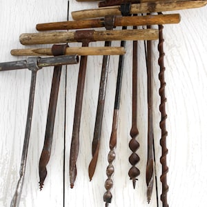 May include: Collection of antique hand drills with wooden handles and metal drill bits. The tools are arranged against a white wooden background, showcasing their aged, rustic appearance. The drills vary in size and design.